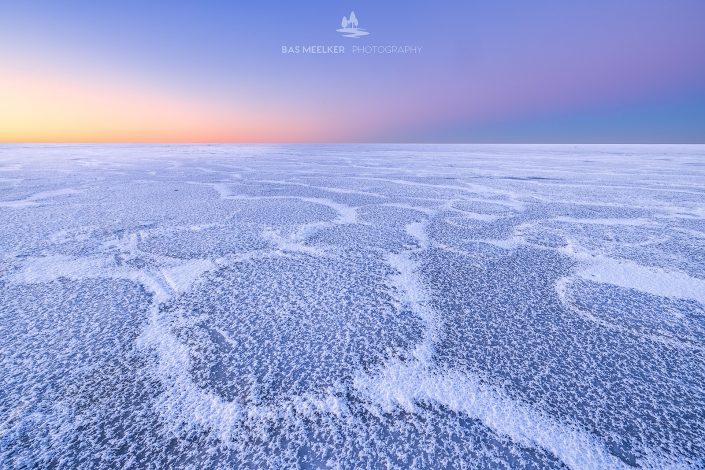 Mooie structuren op een met ijs bedekt IJsselmeer tijdens de winter. Aan de horizon komt de zon langzaam op en geeft een mooie gloed en kleuren aan de lucht.