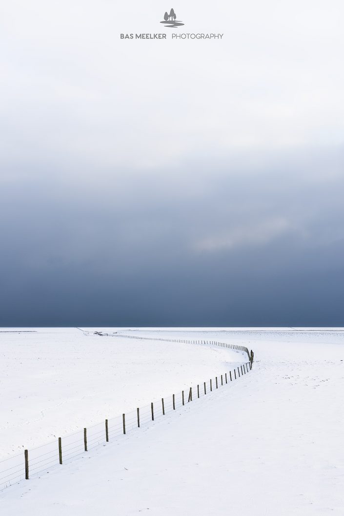 Een leeg winters landschap bedekt met sneeuw in het Nationaal park Lauwersmeer. Aan de horizon drijven donkere sneeuwwolken binnen en geven het landschap een dreigende sfeer. Een minimalistische foto.