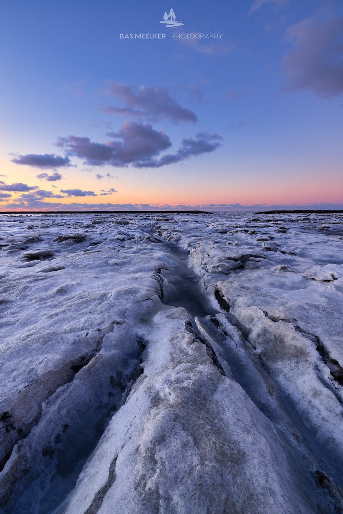 IJs bedekt de Waddenzee in de winter aan de Groningse Waddenkust tijdens zonsondergang. De ondergaande zon geeft een mooi gloed en kleuren aan de lucht.