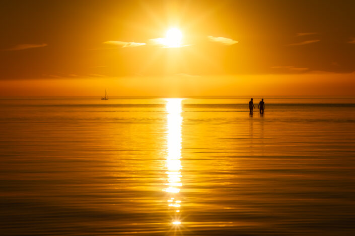 Een zomerse zonsondergang met twee mensen in het water en een zeilboot aan de horizon op een warme dag bij het IJsselmeer.